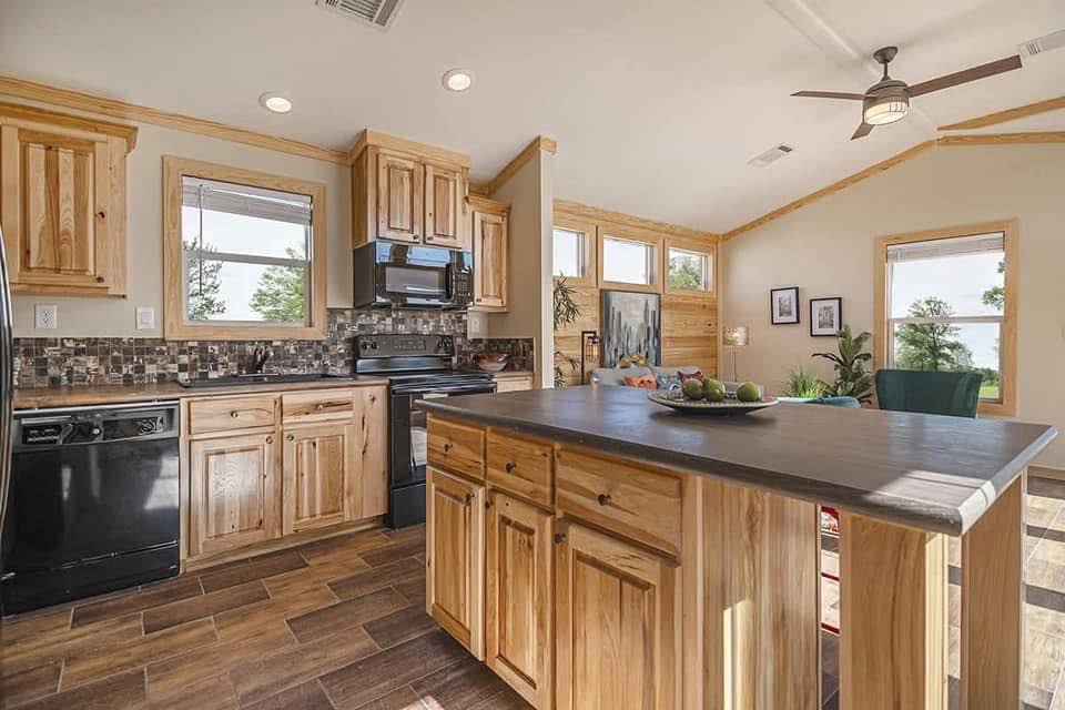A rustic kitchen with wooden cabinets and a large island. Black appliances and a mosaic backsplash add contrast. Sunlight streams through the windows, creating a warm and inviting atmosphere.