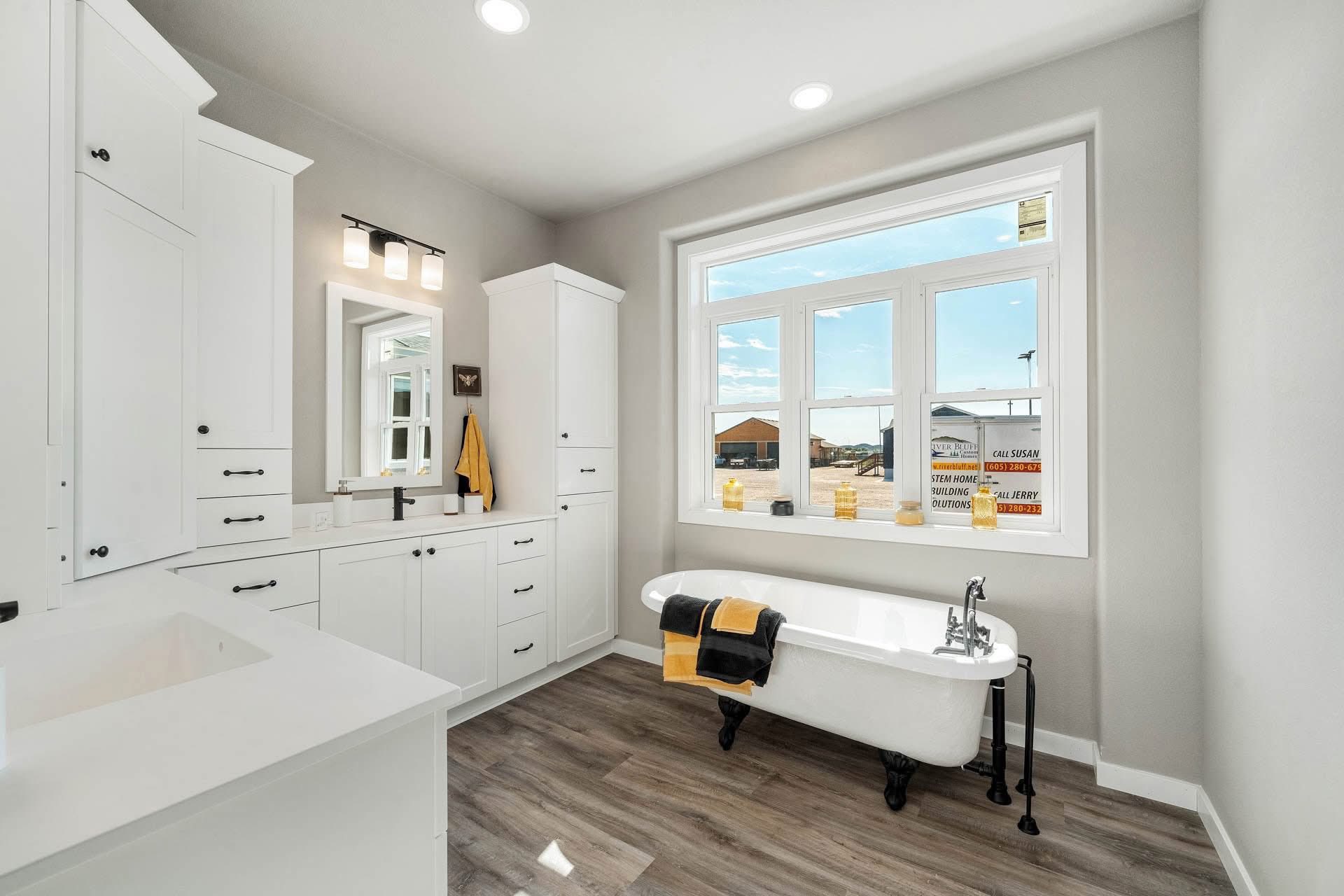 Bright, modern bathroom with white cabinetry and a clawfoot tub. Large window offers natural light. Yellow and black towels add contrast. Warm, inviting atmosphere.