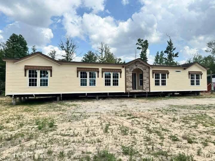 A wide, beige manufactured home with stone accents and multiple large windows sits on a sandy lot. Trees and a partly cloudy sky are in the background.