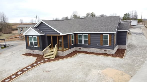 Single-story, dark gray modular home with white trim and brown shutters. A small porch with steps leads to the entrance. Overcast sky above.