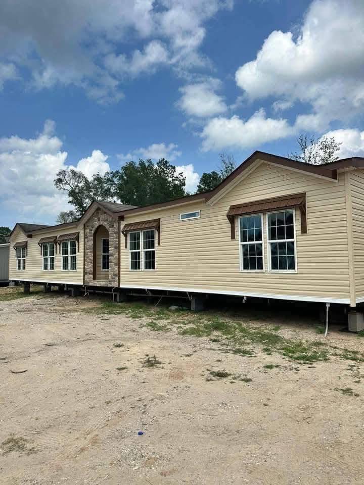 A beige mobile home with multiple large windows and decorative stone entryway sits on a dirt lot under a partly cloudy blue sky, conveying a sense of readiness.