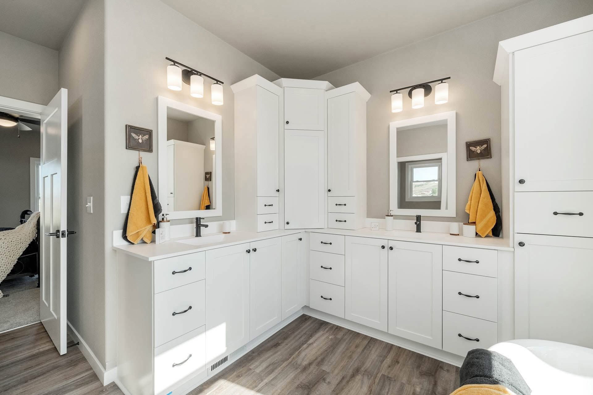 Bright bathroom with white cabinetry, modern fixtures, and dual sinks. Yellow and black towels hang beside mirrors beneath warm lighting.