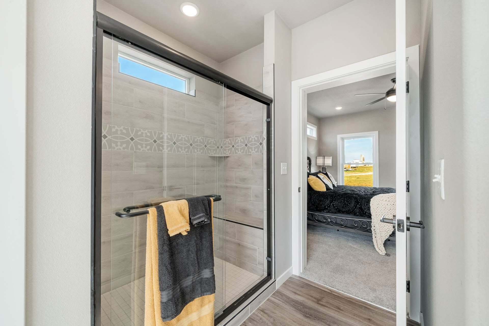 Modern bathroom with a glass-door shower, gray tile, and decorative trim. Yellow and black towels hang on the rail. Open door leads to a cozy bedroom.