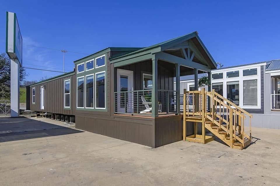 Modern modular home with a wooden porch, green accents, and large windows under a clear blue sky. A wooden staircase leads to the entrance.