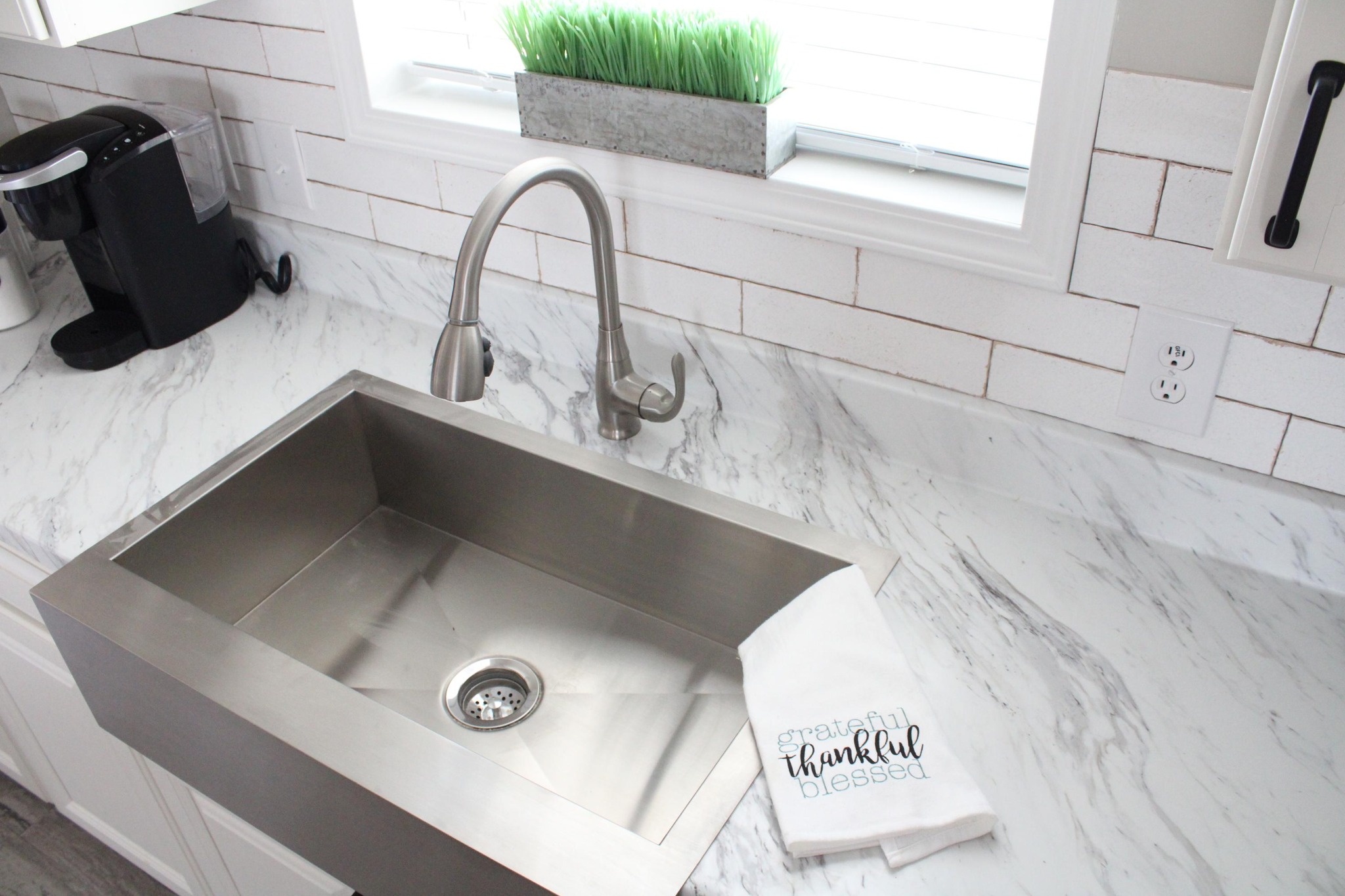 Modern kitchen with a stainless steel sink, marble countertop, and a black coffee maker. A towel reads "grateful, thankful, blessed." A window above adds brightness.