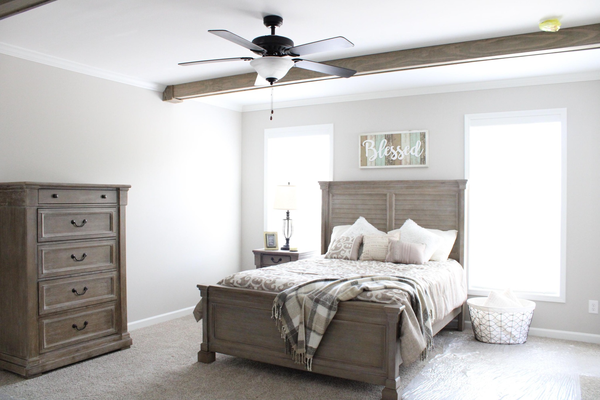 Cozy bedroom with a wooden bed, plaid blanket, and pillows. "Blessed" sign above bed. Natural light from two windows; dresser and ceiling fan visible.