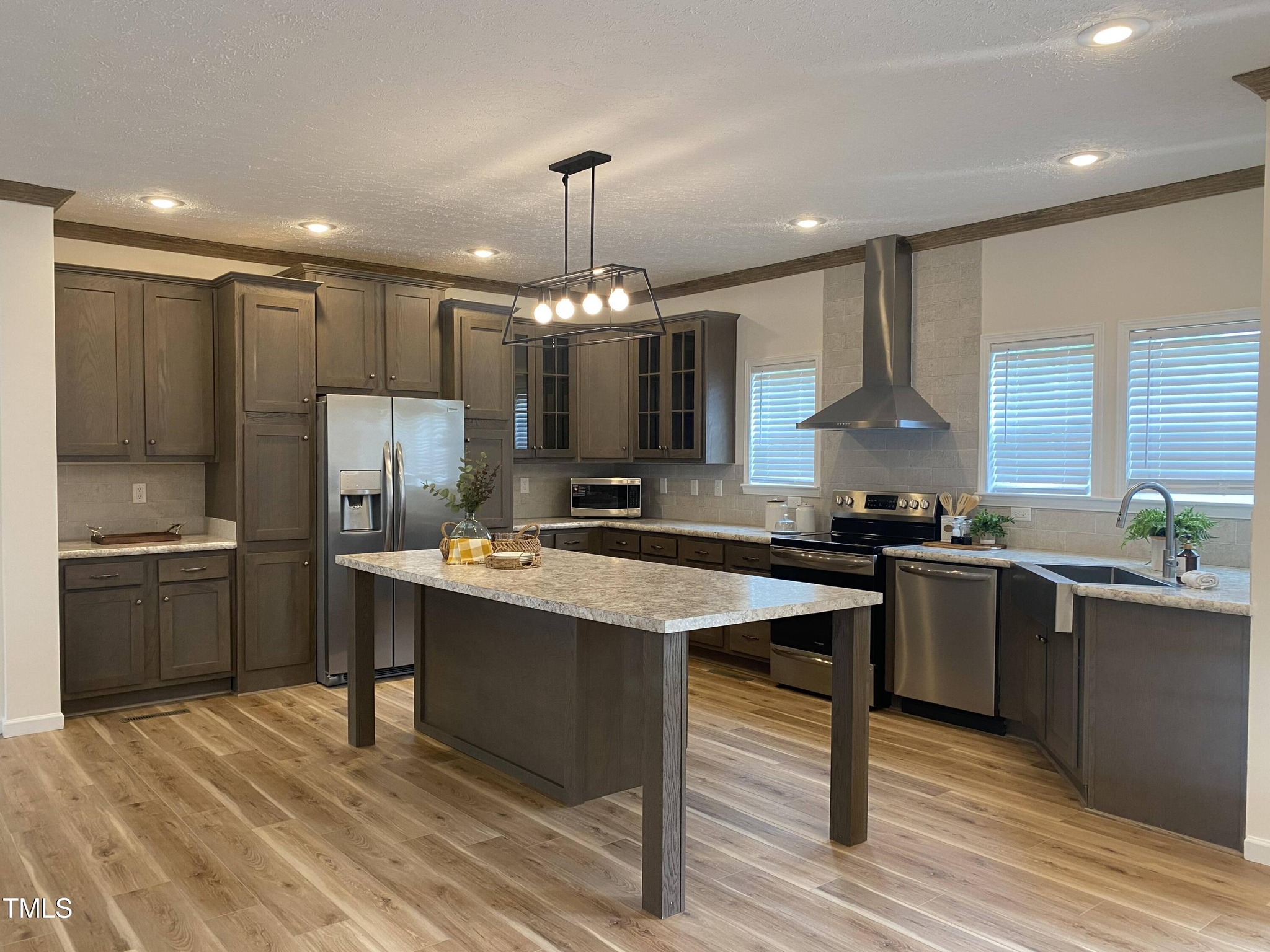 Modern kitchen with gray cabinetry, stainless steel appliances, and a large island. Wood flooring and large windows create a bright, airy feel.