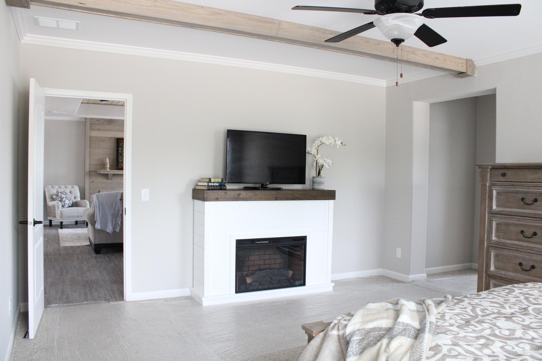 Modern bedroom with beige walls features a white fireplace with a TV on top, a ceiling fan, and visible hallway. Cozy and neutral tone.