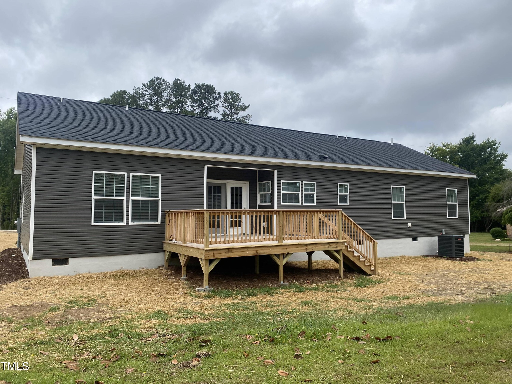 A modern gray house with white trim and multiple windows, featuring a raised wooden deck with railing. Overcast sky and trees in the background.