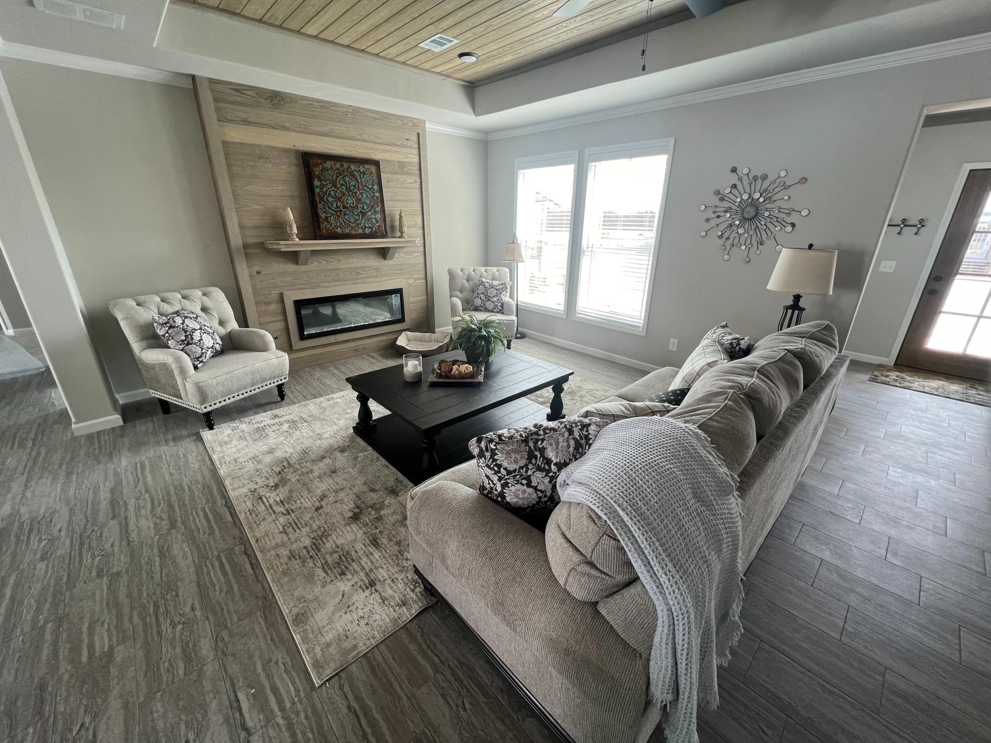 Cozy living room with beige sofa, patterned cushions, and armchairs. Features a wood fireplace, abstract wall art, and large windows. Neutral tones, inviting atmosphere.