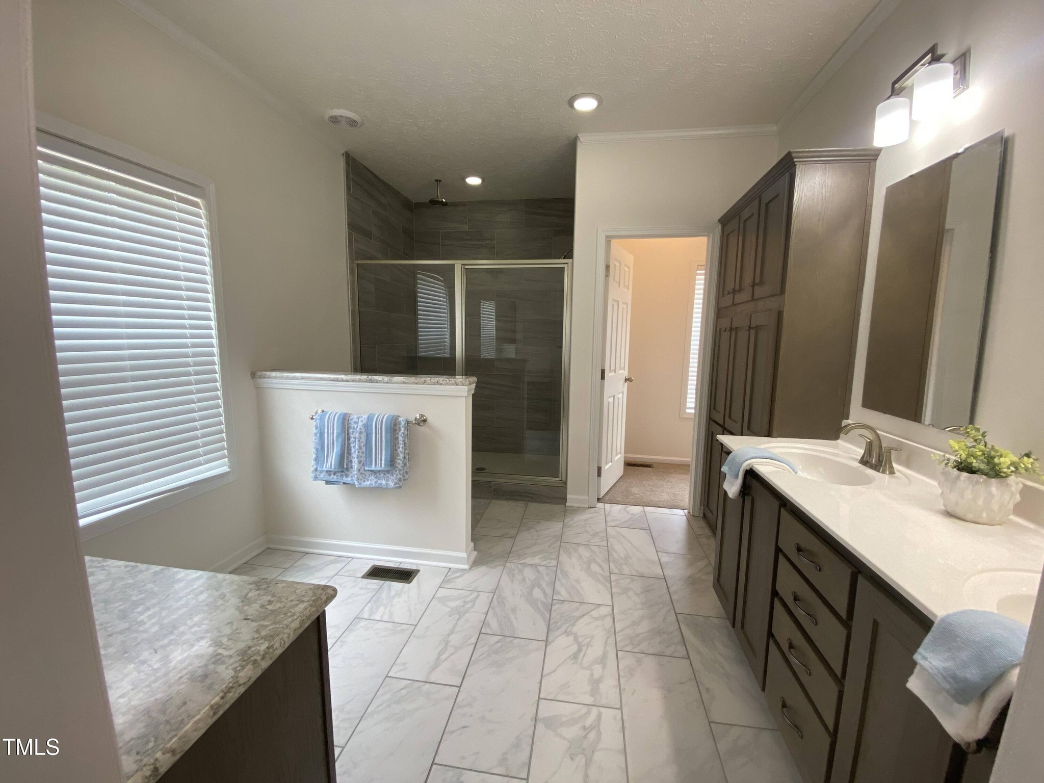 Spacious bathroom with grey tiles and white walls. Features a double sink vanity, large mirror, glass shower, and blue towels, conveying elegance.