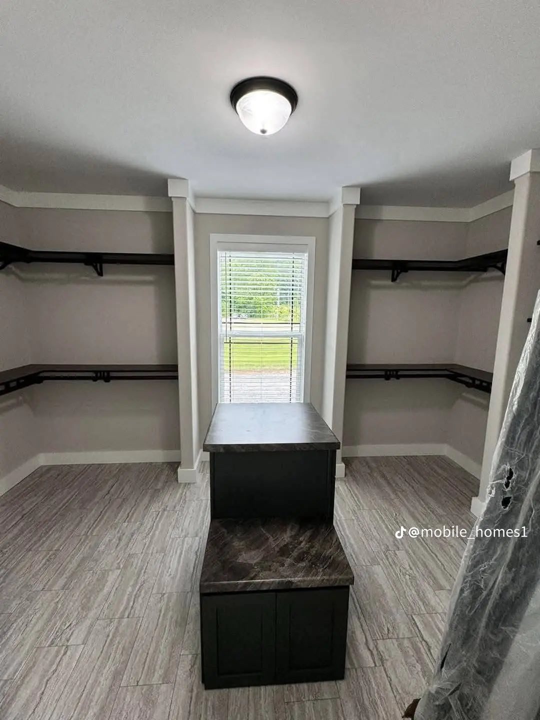 Spacious walk-in closet with light wood floors, beige walls, and dark wood shelving. A window with blinds lets in natural light. Mood is bright and organized.