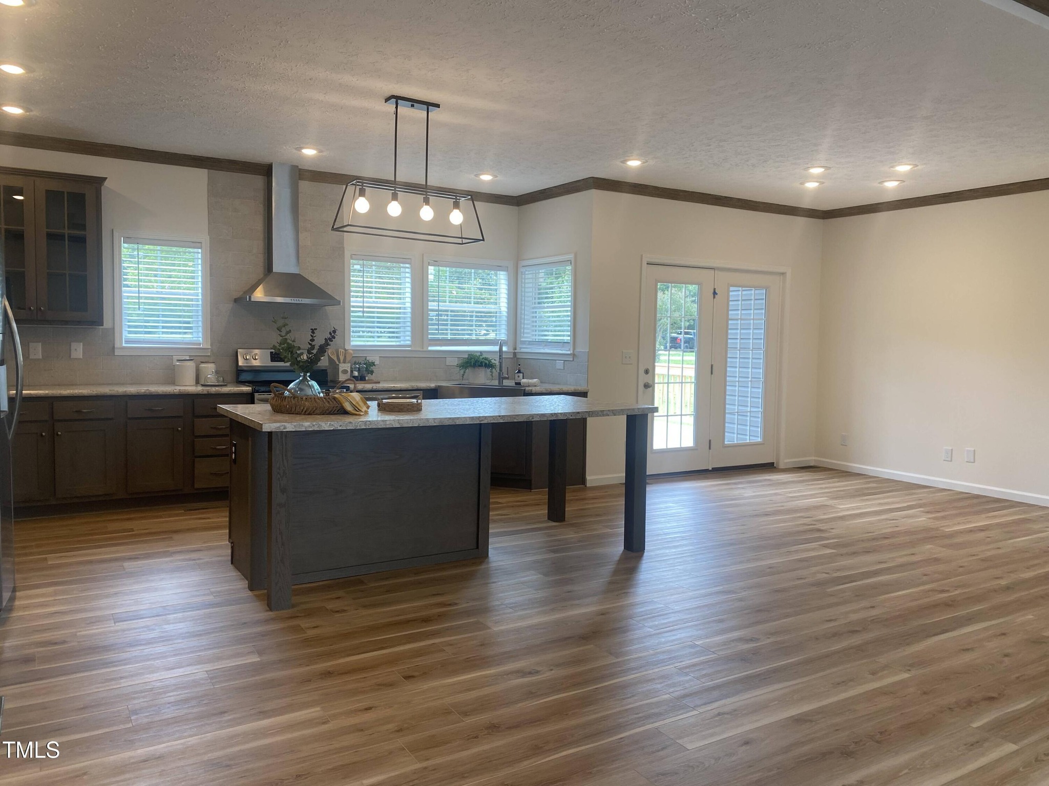 Modern kitchen with wooden floors and dark cabinets. A large island with a pendant light fixture is central. Windows let in ample natural light, creating a warm, inviting atmosphere.