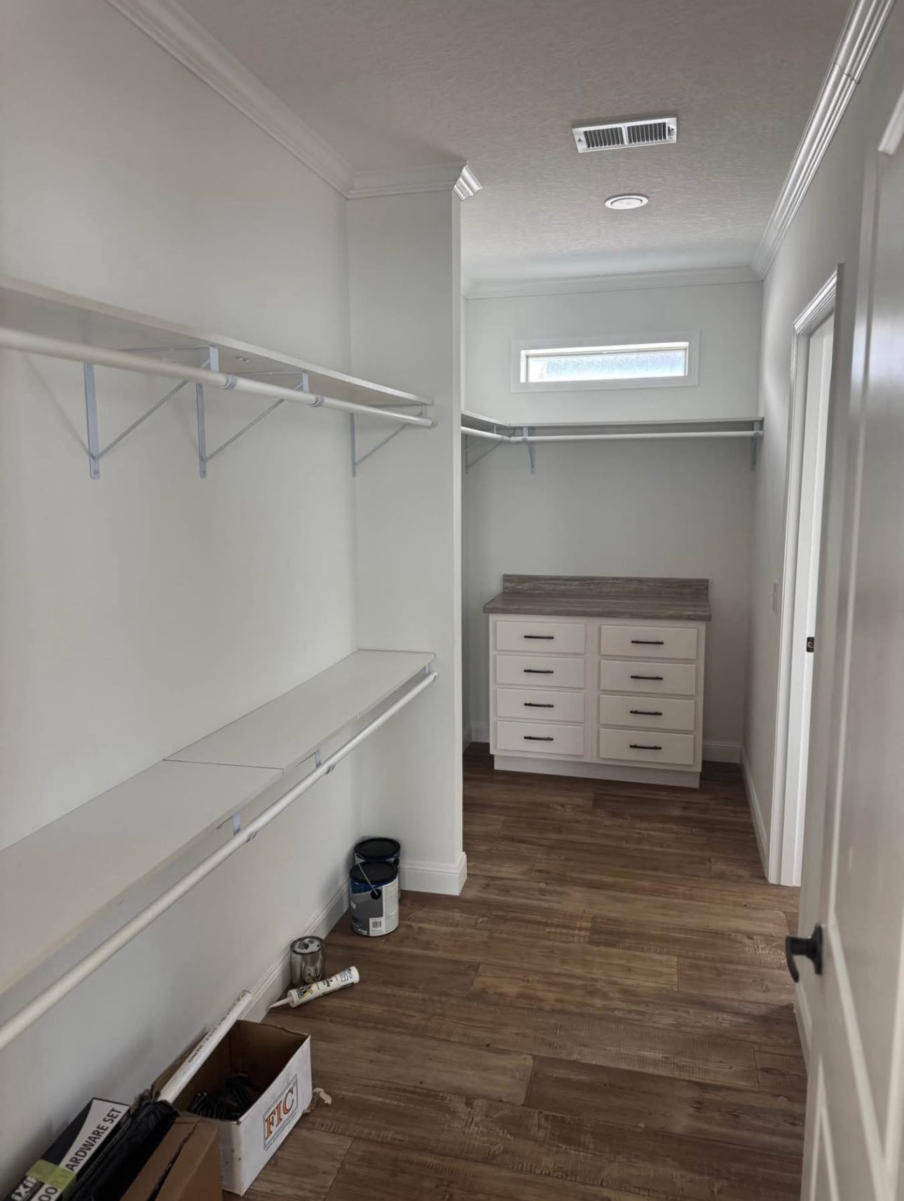 Narrow walk-in closet with wooden floor, white walls, shelving, and a dresser with six drawers. A small window provides natural light.