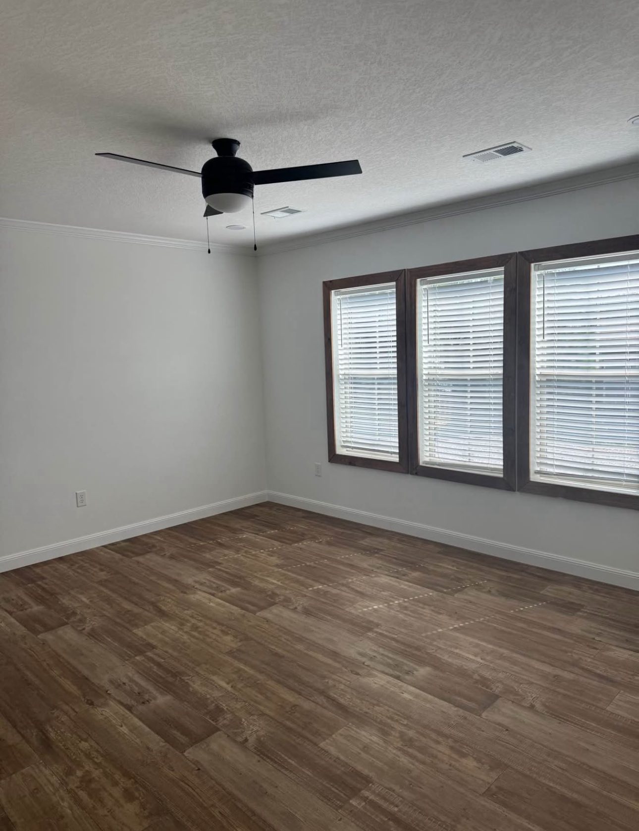 A vacant room with white walls and wood flooring. Three large windows with blinds allow light in. A sleek black ceiling fan is centrally positioned.
