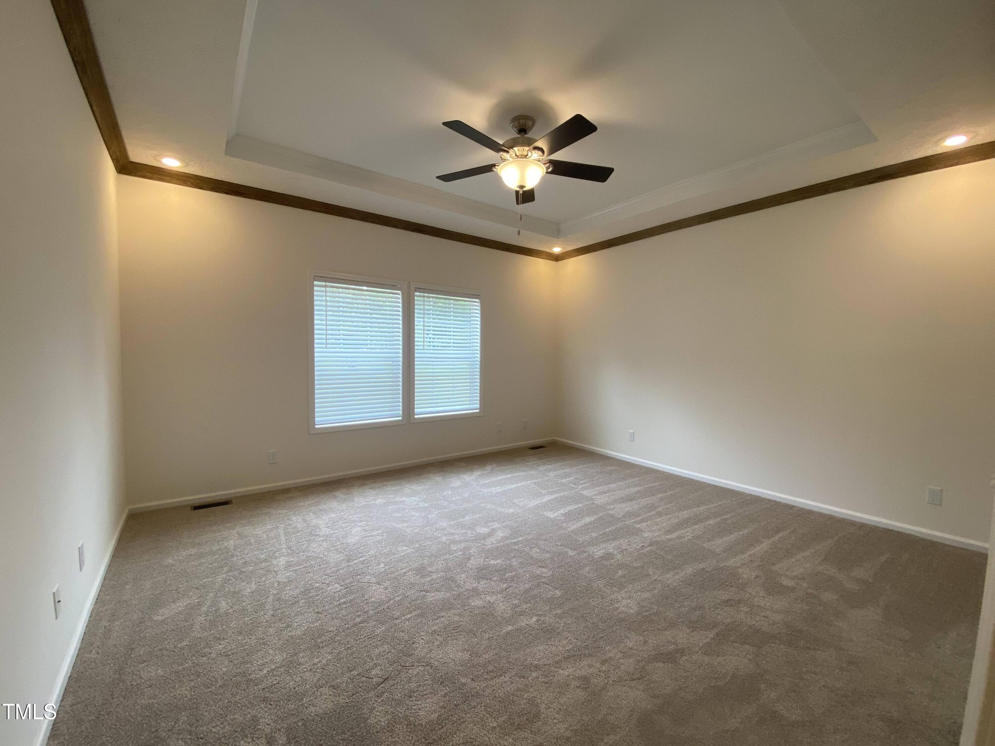 Spacious empty room with beige carpet and cream walls. Features a tray ceiling with recessed lights and a central ceiling fan. Two large windows provide natural light.