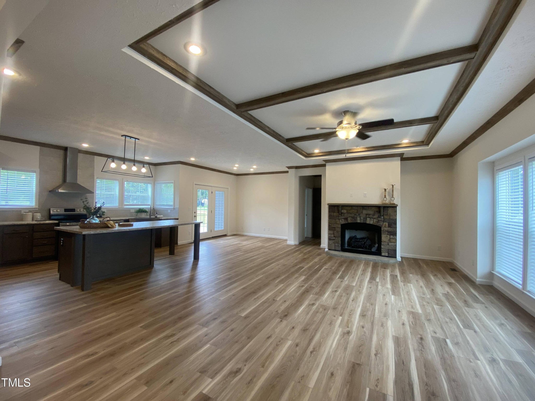 Spacious open-plan room with light wood flooring, featuring a modern kitchen with a dark island, a stone fireplace, and a ceiling fan, creating a cozy and inviting atmosphere.