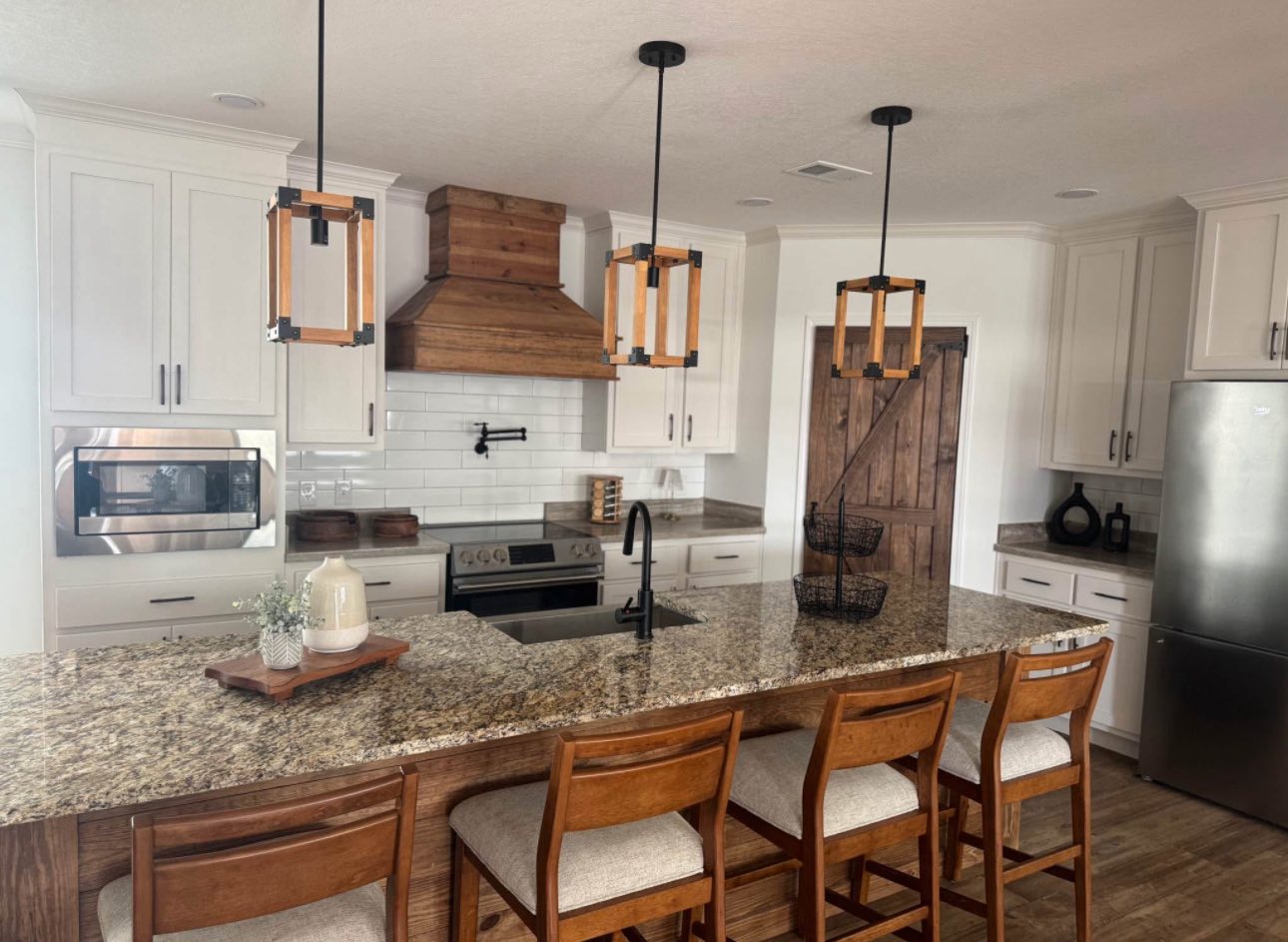 Modern kitchen with white cabinets, a wood range hood, and granite countertops. Three pendant lights hang over an island with wooden stools, creating a warm, inviting atmosphere.