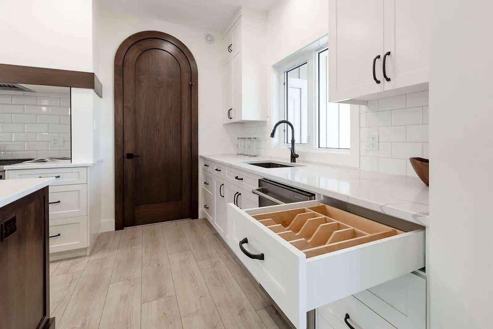 Modern kitchen with light wood floor, white cabinets, and open drawer with dividers. Features a dark wood arched door and black fixtures for contrast.