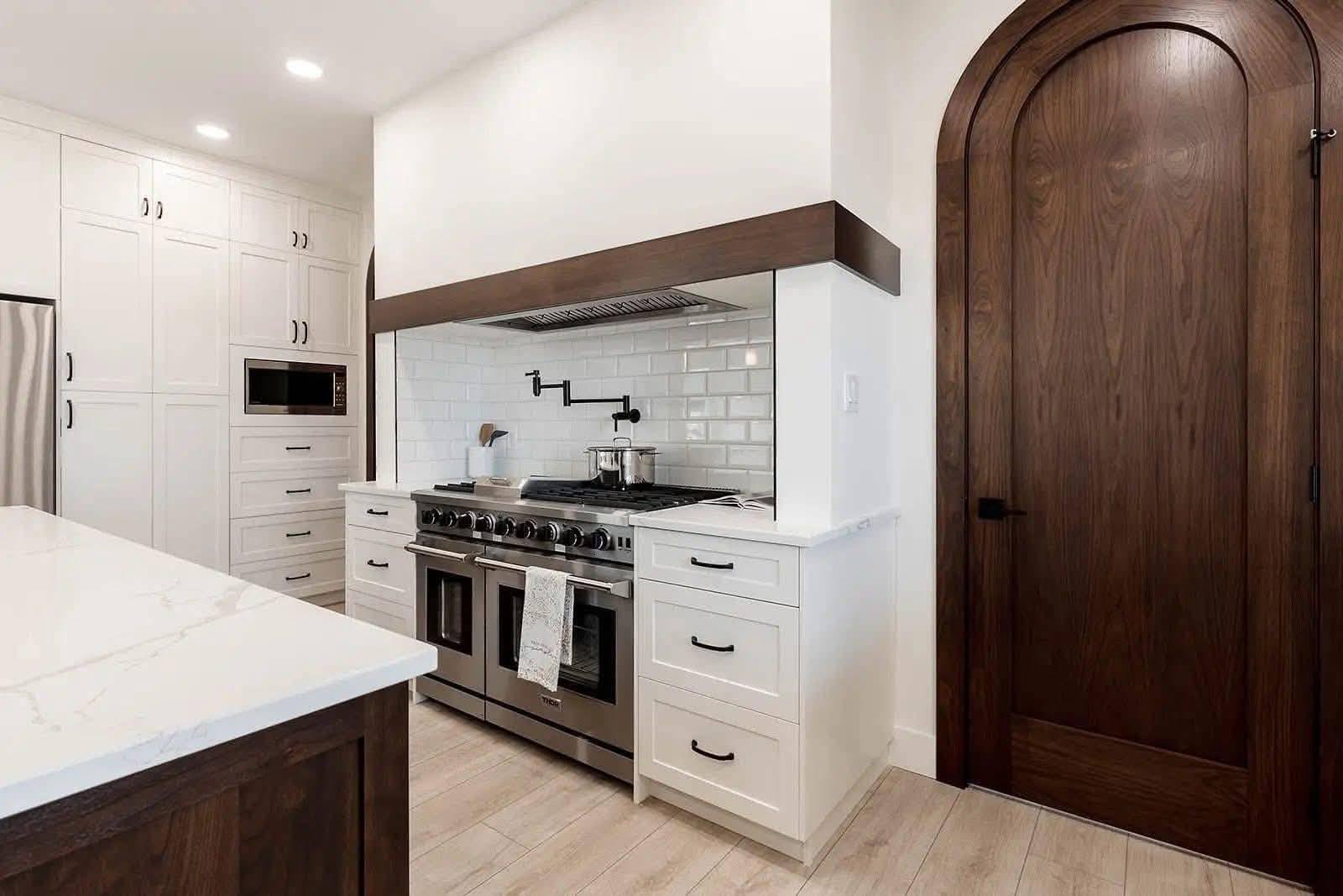 Modern kitchen with white cabinetry, stainless steel appliances, and wood accents. Large arched wooden door adds warmth and elegance. Bright, clean tone.
