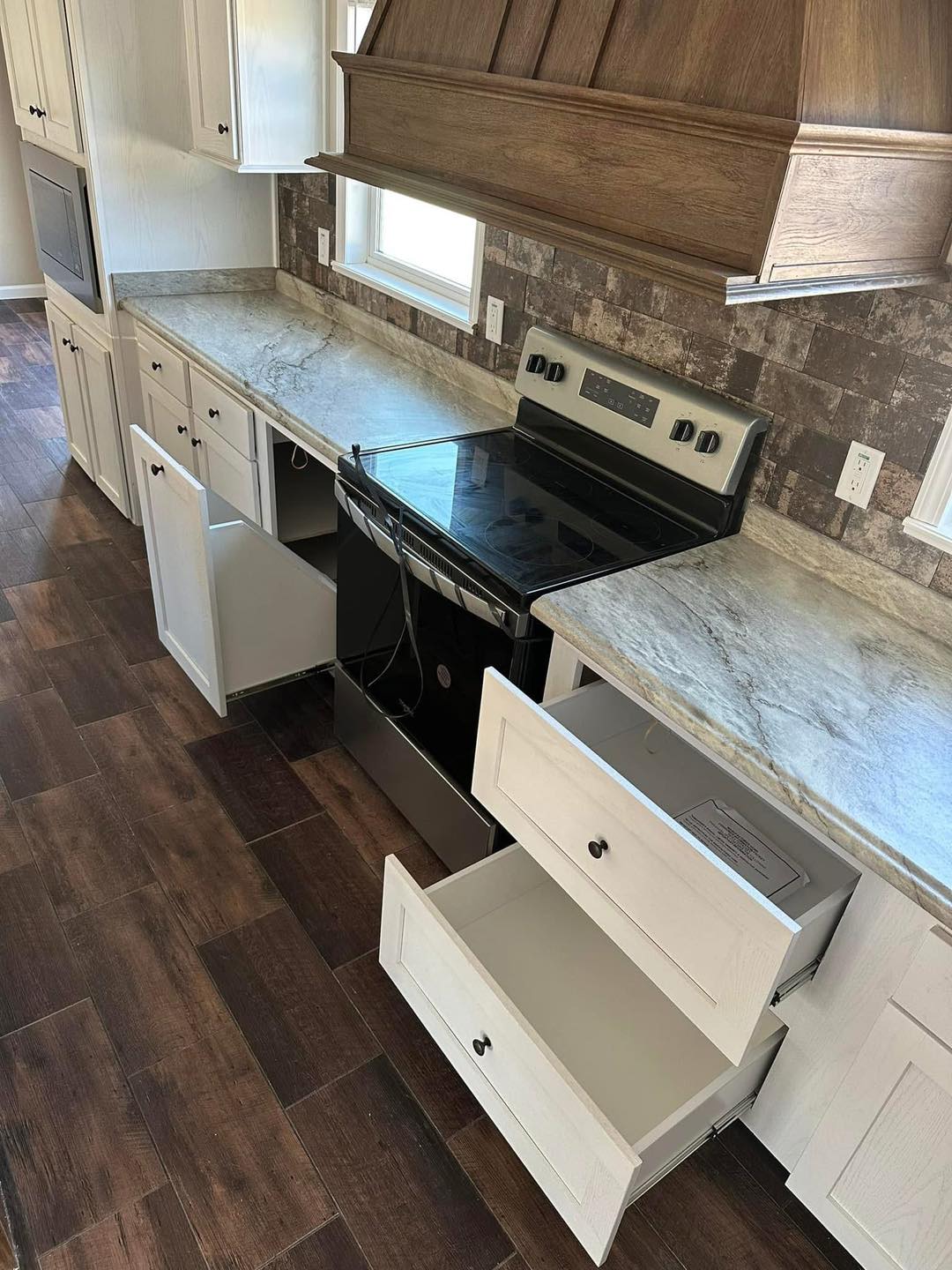 Kitchen with brown wood floors, white cabinets, and a marble-look countertop. Open drawers show storage; an electric stove is centrally placed.