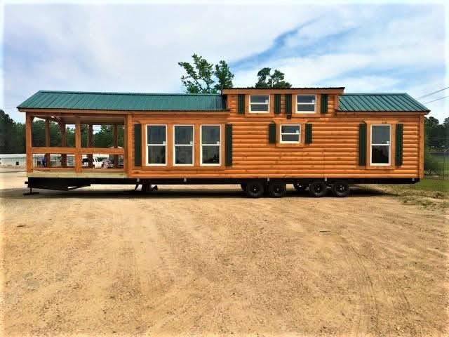Log cabin-style mobile home with green metal roof on wheels, parked on dirt. Large windows and porch evoke a rustic yet cozy feel.