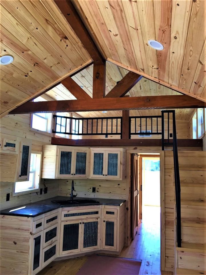 Cozy wooden tiny house interior with a loft. Features a peaked ceiling, rustic cabinets, black metal railing, and natural light, creating a warm atmosphere.