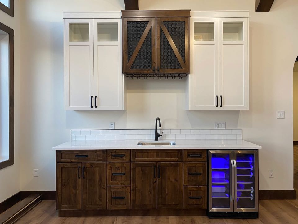 Modern kitchen bar area with white upper cabinets and dark wood lower cabinets. Features a small sink, dark faucet, and a lit wine cooler.
