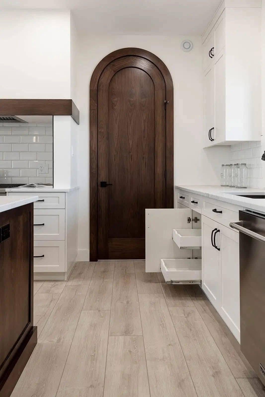 Sleek kitchen with light wood floors, white cabinets, dark wood accents, arched wooden door, tile backsplash, open drawers, and clean lines.