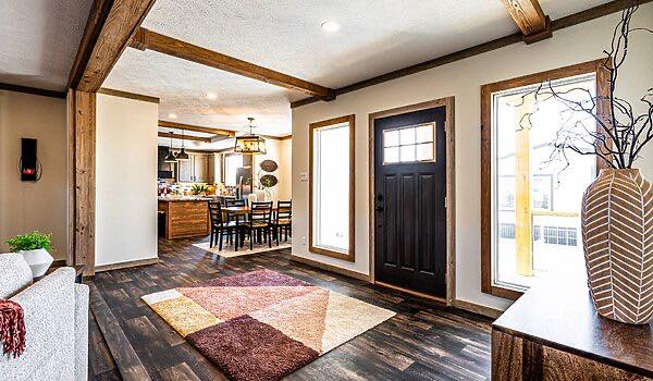 Warm, inviting living room with dark wood beams and flooring, a geometric rug, and large windows. Stylish dining area in the background, conveying coziness.