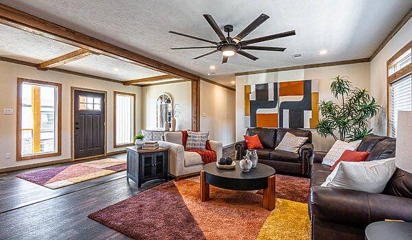 Warm, modern living room with dark wood floors, large ceiling fan, abstract art, two sofas with colorful pillows, and geometric-patterned rugs.