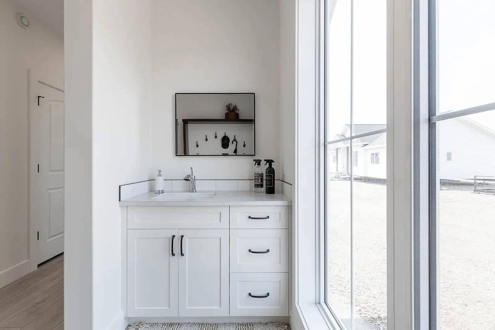 Bright, minimalist bathroom with a white vanity and black handles. A rectangular mirror hangs above, reflecting a shelf with decor. Large window to the right.