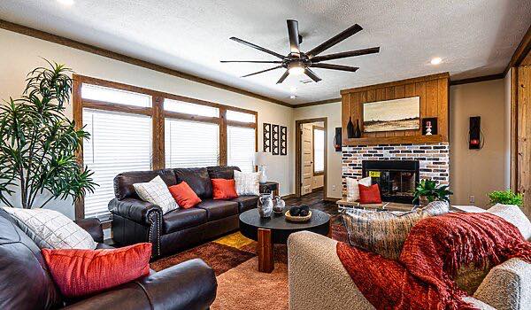 Stylish living room with dark leather sofa and beige armchairs, red and white cushions, a modern ceiling fan, brick fireplace, and large windows. Cozy and inviting.