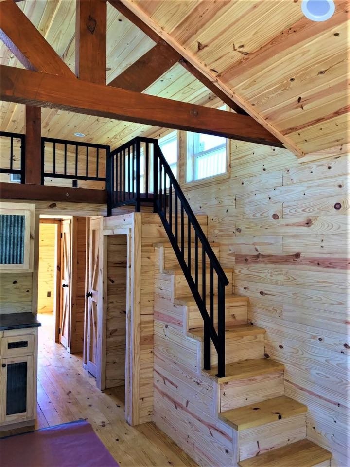 Modern tiny home interior with wooden walls and ceiling, featuring a compact staircase with black railings leading to a loft. Warm, cozy atmosphere.