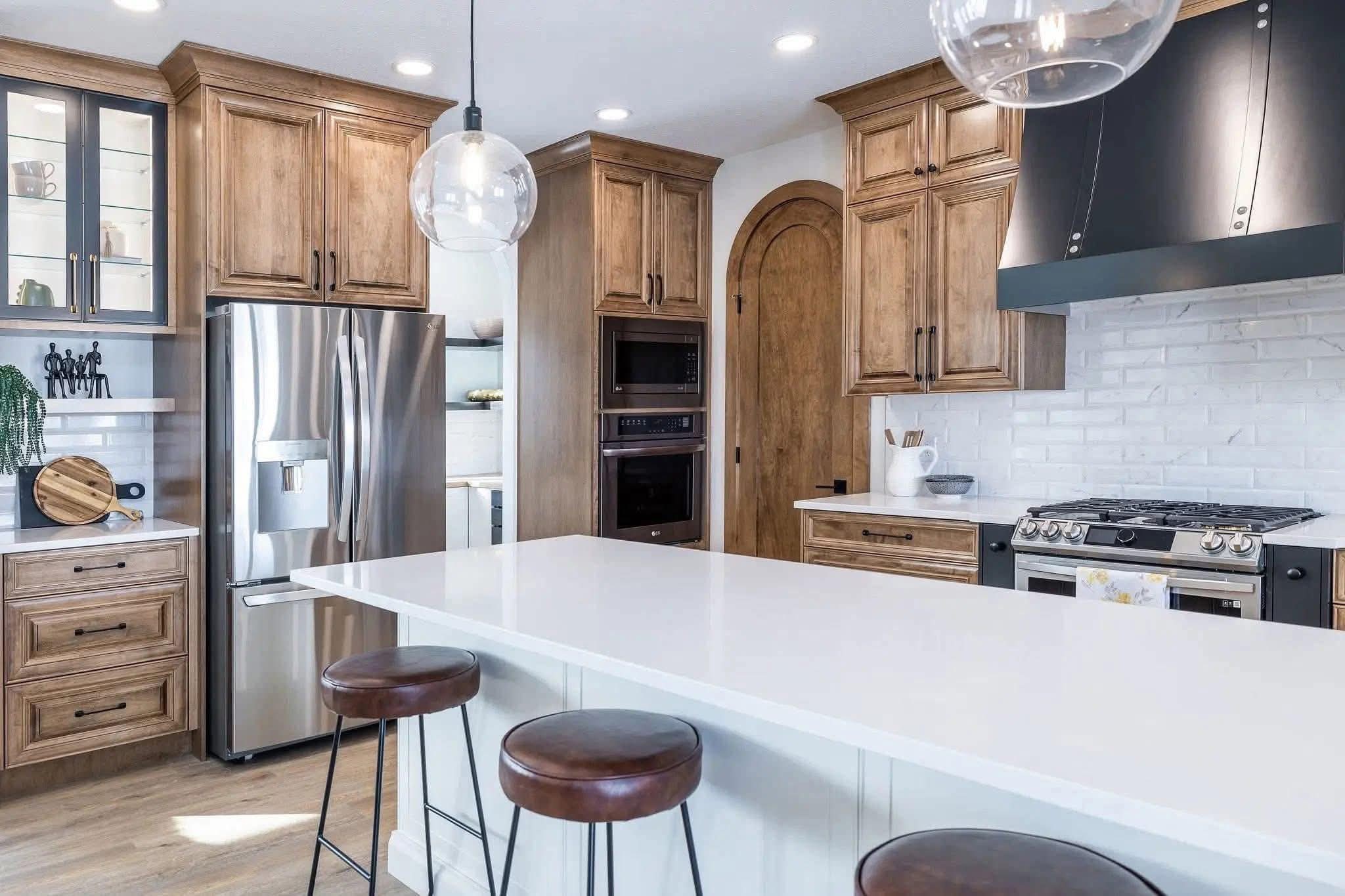 Modern kitchen with wooden cabinets, stainless steel fridge, and black range hood. White island with bar stools and pendant lighting. Bright, airy feel.
