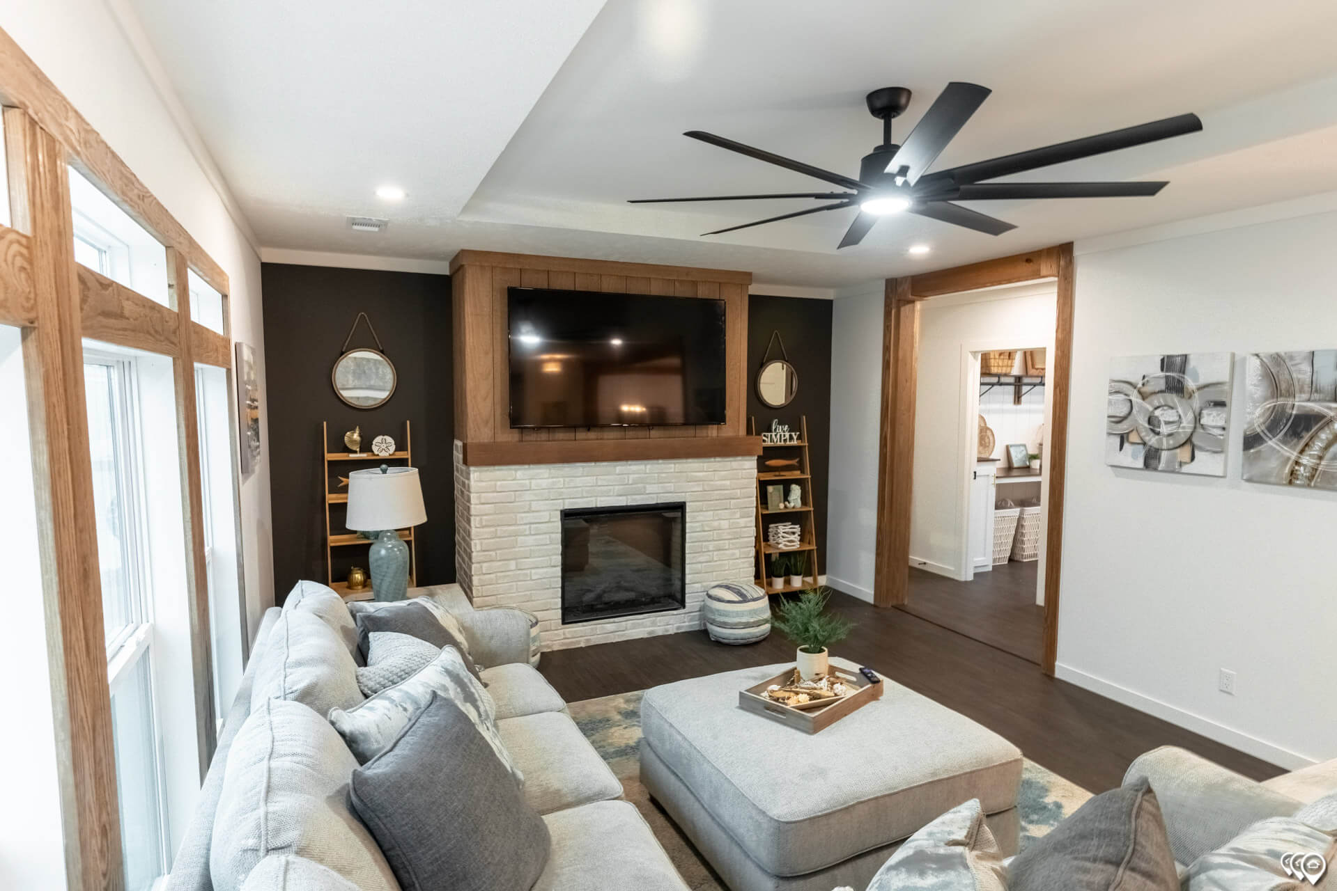 Modern living room with a large gray sectional sofa, centered around a brick fireplace with a mounted TV. The room features warm wood accents, a black ceiling fan, and decorative shelves, creating a cozy, inviting atmosphere.