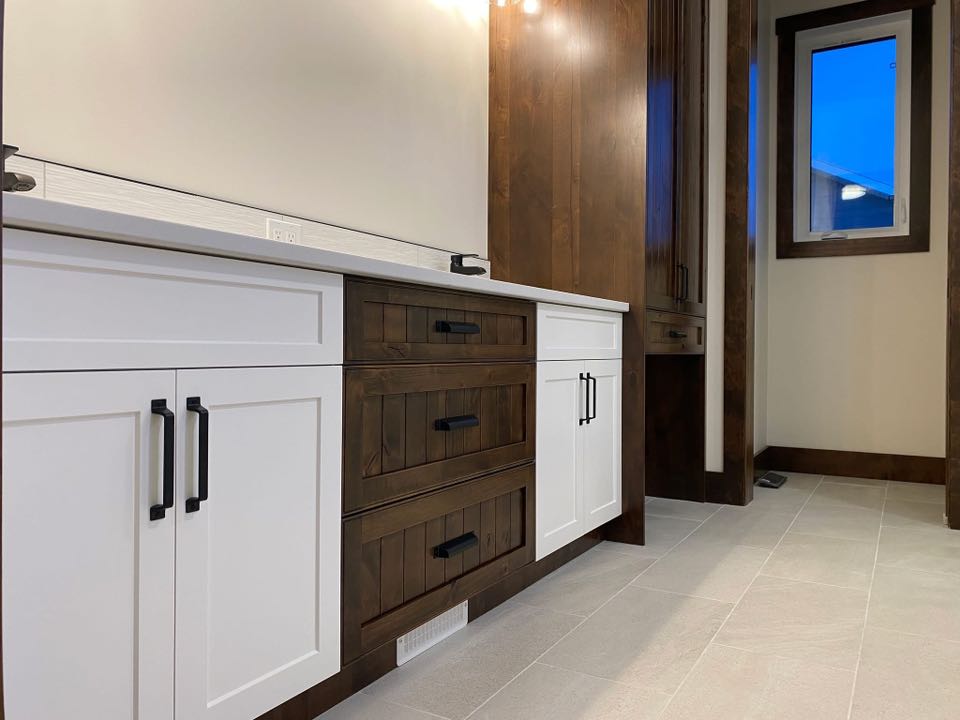 Modern kitchen interior with white and dark wood cabinets, black handles, and beige tile flooring. A window shows a twilight sky, creating a warm ambiance.