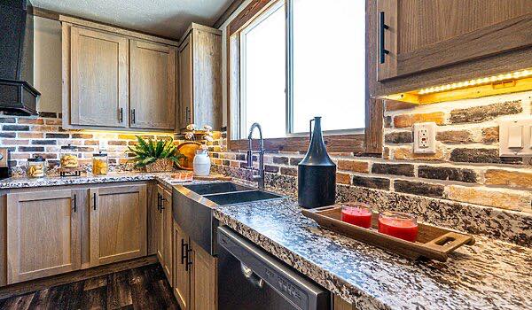 Warm kitchen with wooden cabinets, brick backsplash, and granite countertops. A window illuminates the space. Red candles and decorative items add coziness.