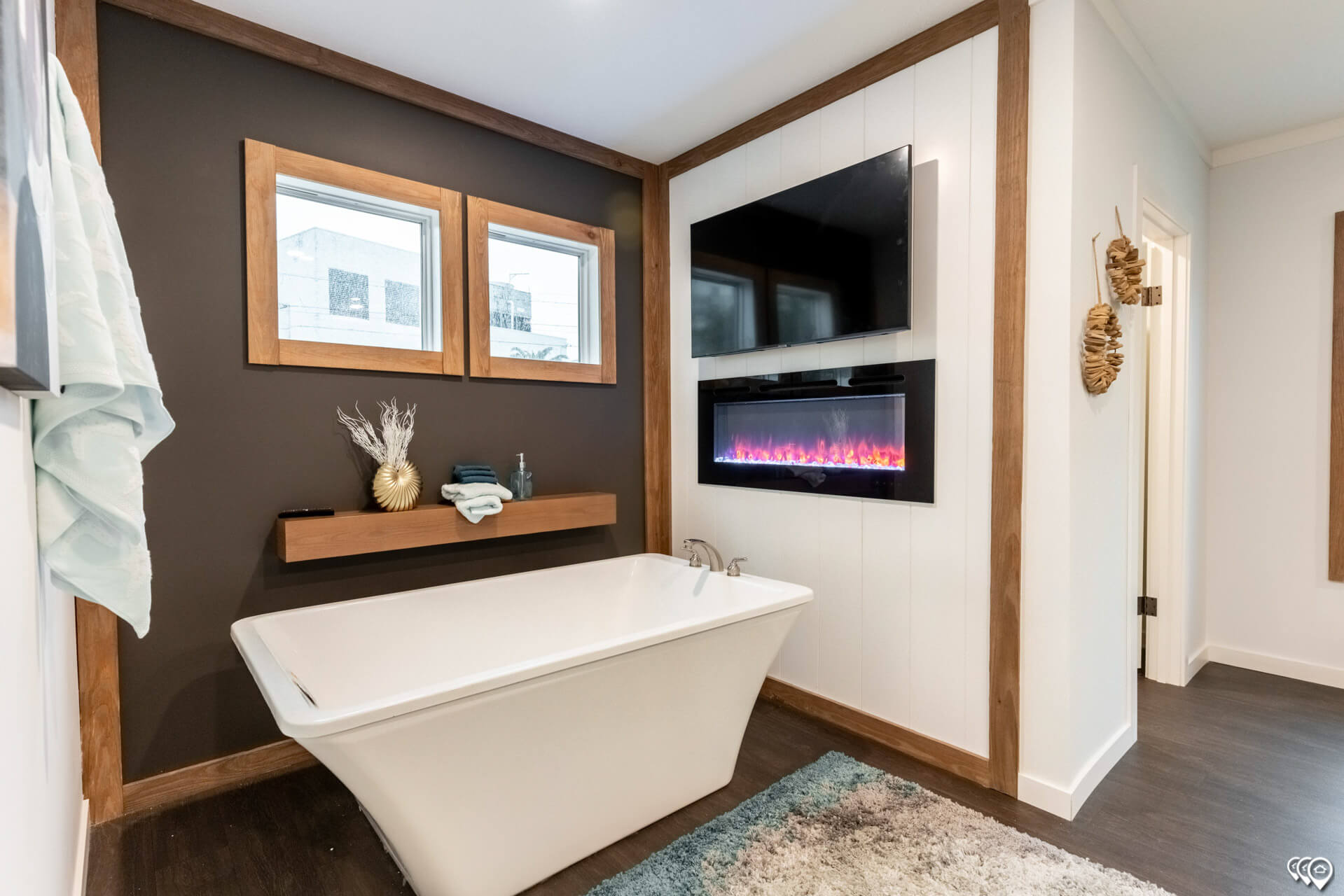 Modern bathroom with a white freestanding tub, dark accent wall, and two large windows. Mounted TV and electric fireplace add warmth and luxury.