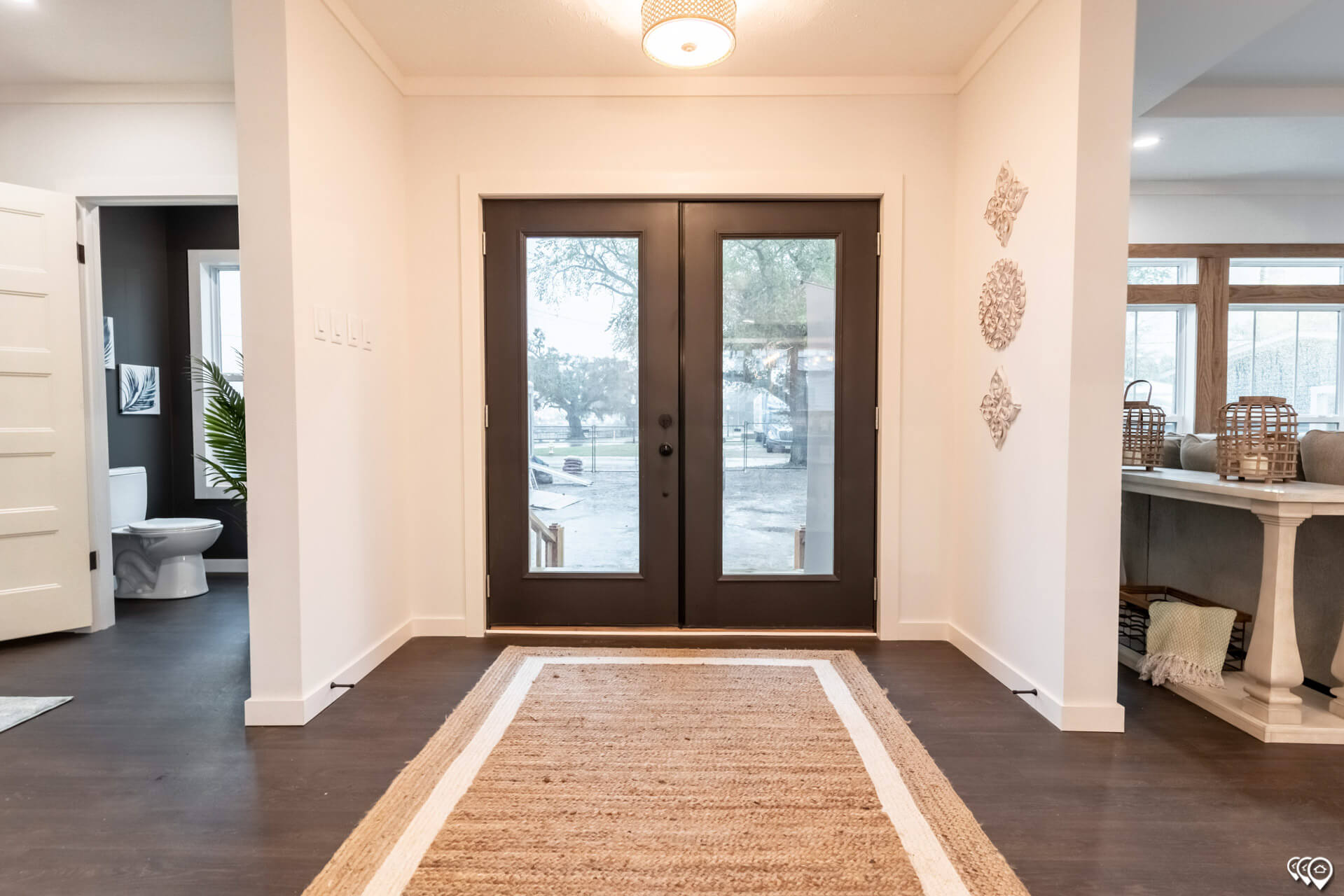 Spacious entryway with dark glass double doors. A woven rug on wooden floors, art pieces on white walls, and a glimpse of a bathroom highlight the modern decor.