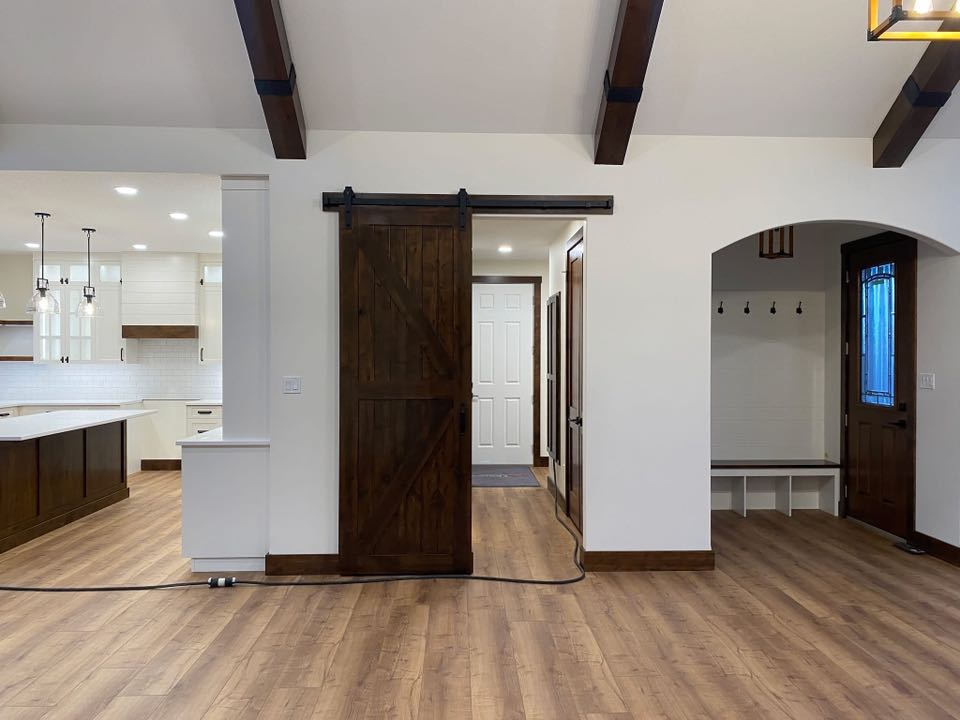 Spacious, modern interior with wooden barn door, light wood flooring, and white walls. Kitchen with hanging lights on the left; cozy nook and entryway on the right.