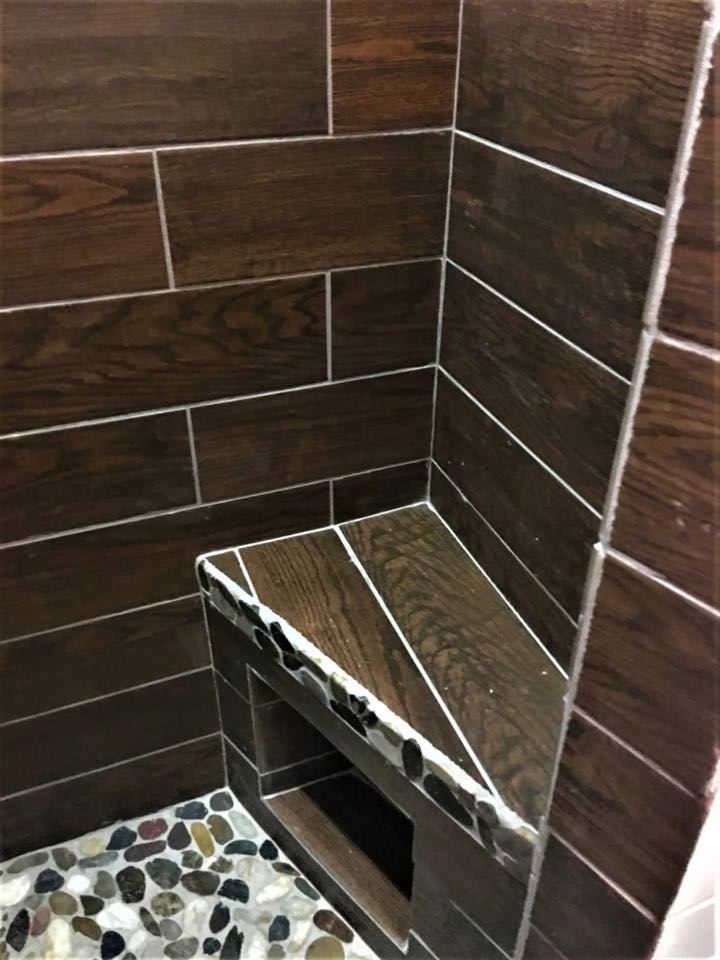 Corner of a bathroom shower with dark wood-look tiles and a small triangular seat. Pebble-style flooring adds texture, creating a rustic, cozy feel.