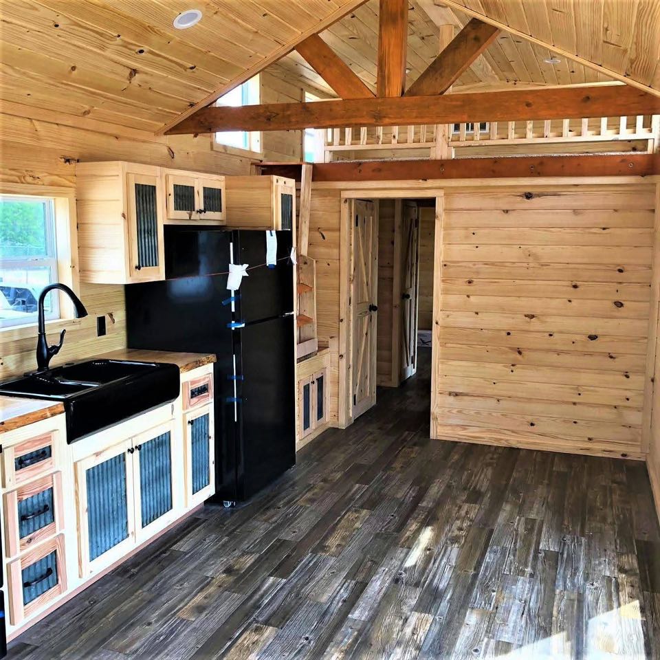 Interior of a wooden tiny house with vaulted ceilings. Features include a black fridge, farmhouse sink, and rustic cabinets, creating a cozy atmosphere.