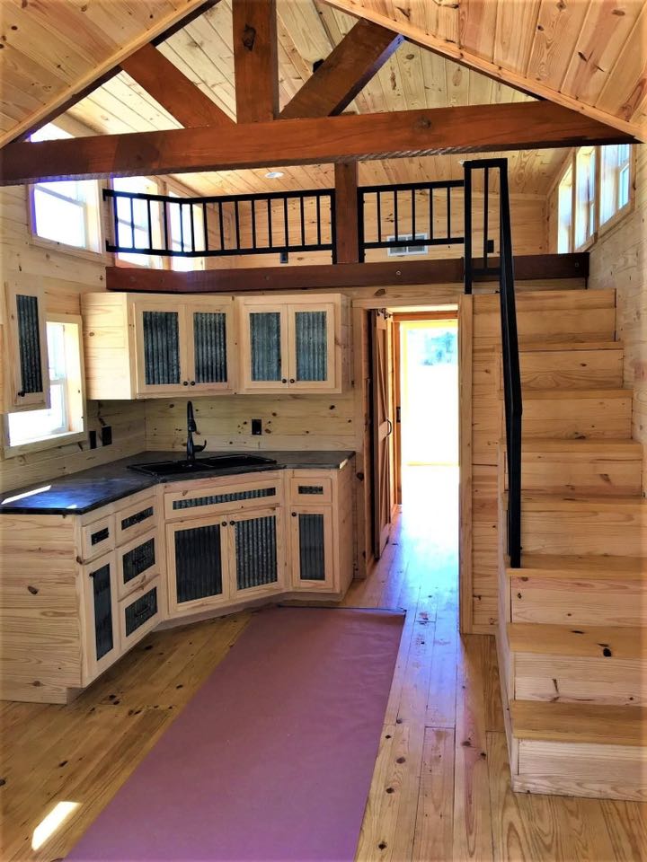 Interior of a cozy wooden tiny house featuring a corner kitchen with rustic cabinets. A loft with railing is accessed by a wooden staircase, creating a warm and inviting atmosphere.