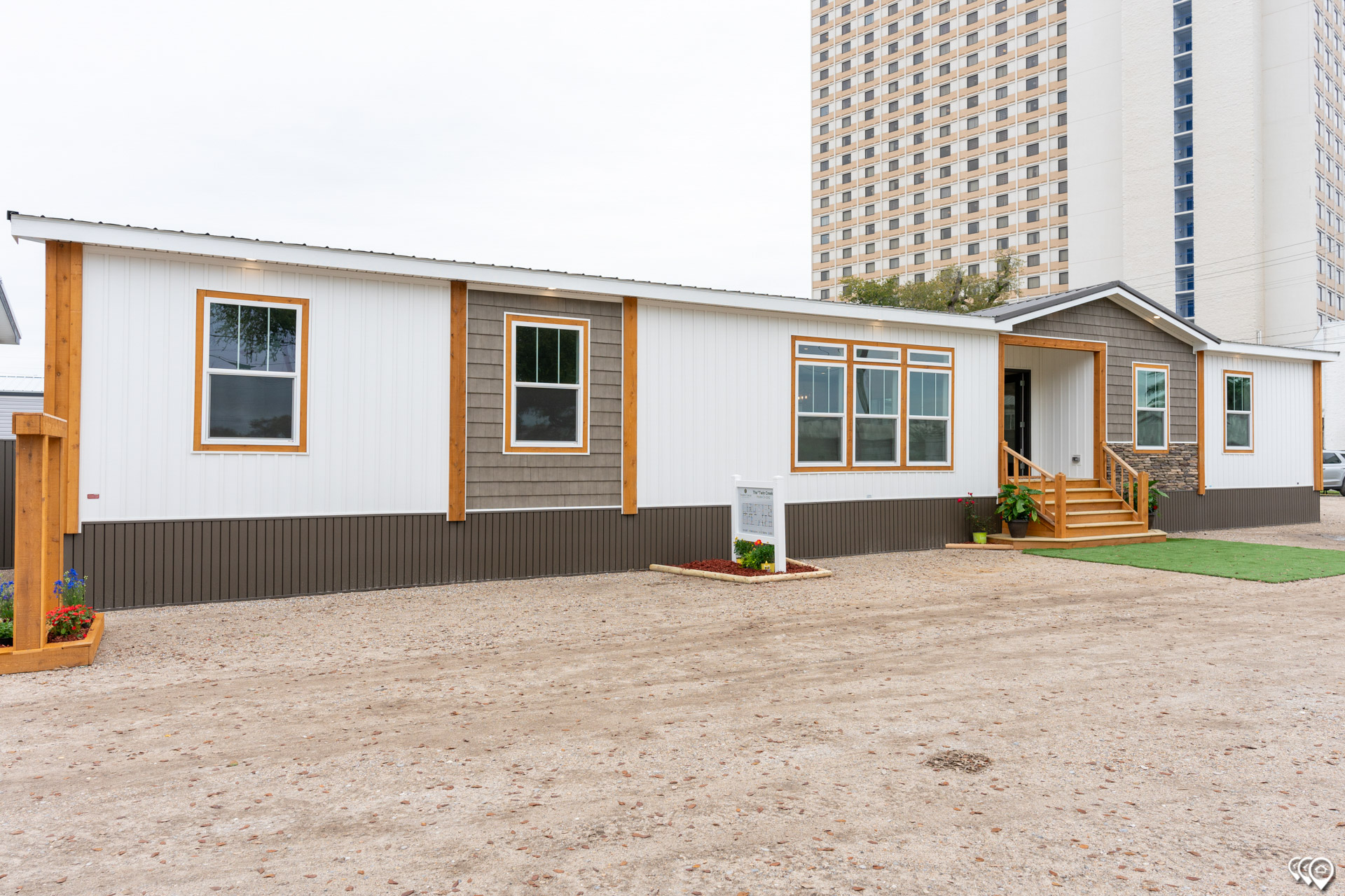 Modern modular home with white and gray siding, wooden accents, surrounded by a gravel path. A tall building looms in the background, contrasting styles.