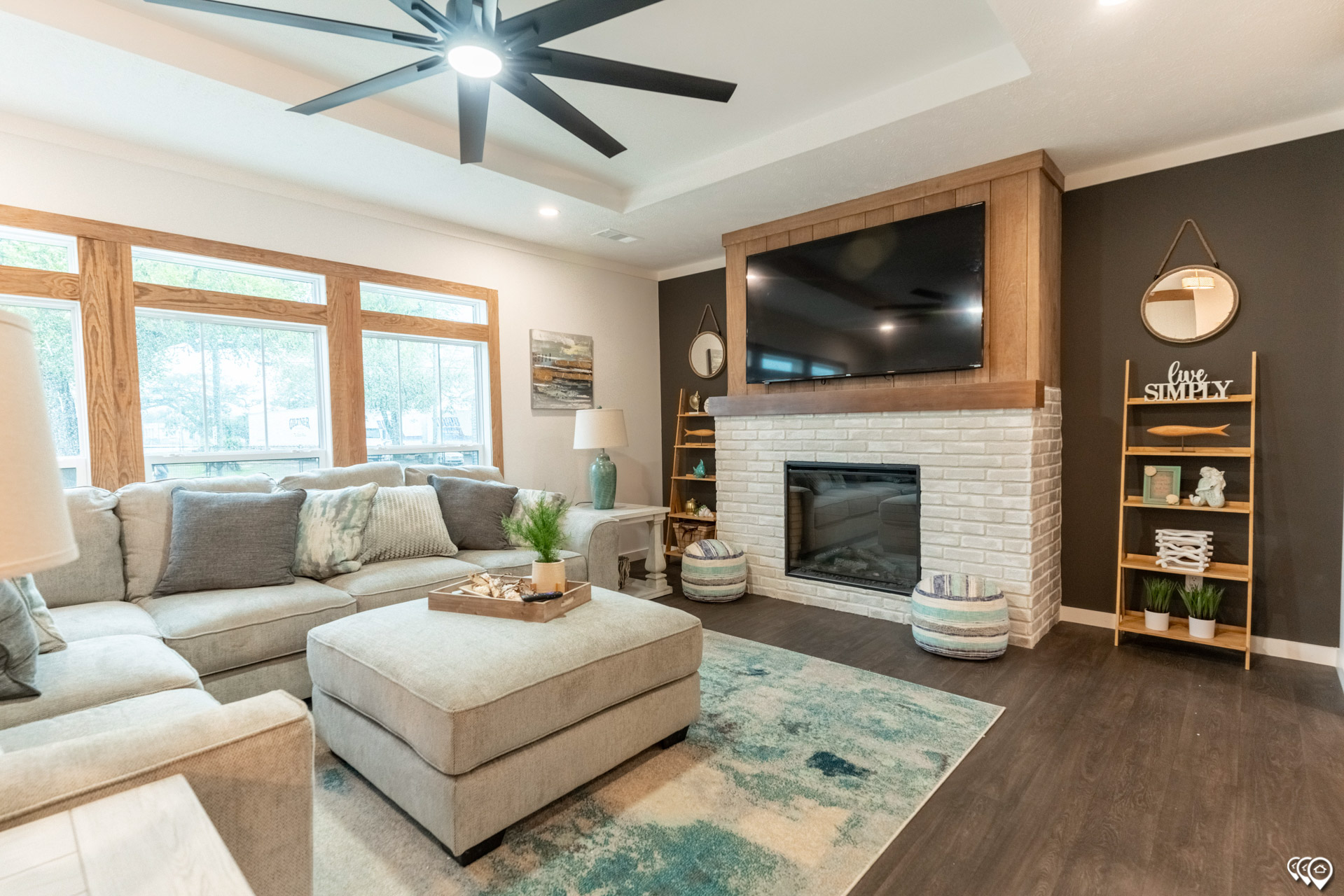A cozy living room with a large gray sectional sofa and a matching ottoman on a blue and beige rug. A brick fireplace is topped with a flat-screen TV. Wooden shelves with decor flank the fireplace, while large windows and a ceiling fan enhance the space's brightness and comfort.