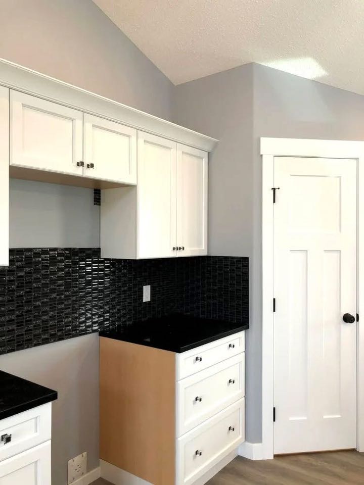 Modern kitchen corner with white cabinets, black countertops, and a textured black tile backsplash. Light gray walls and a paneled white door.