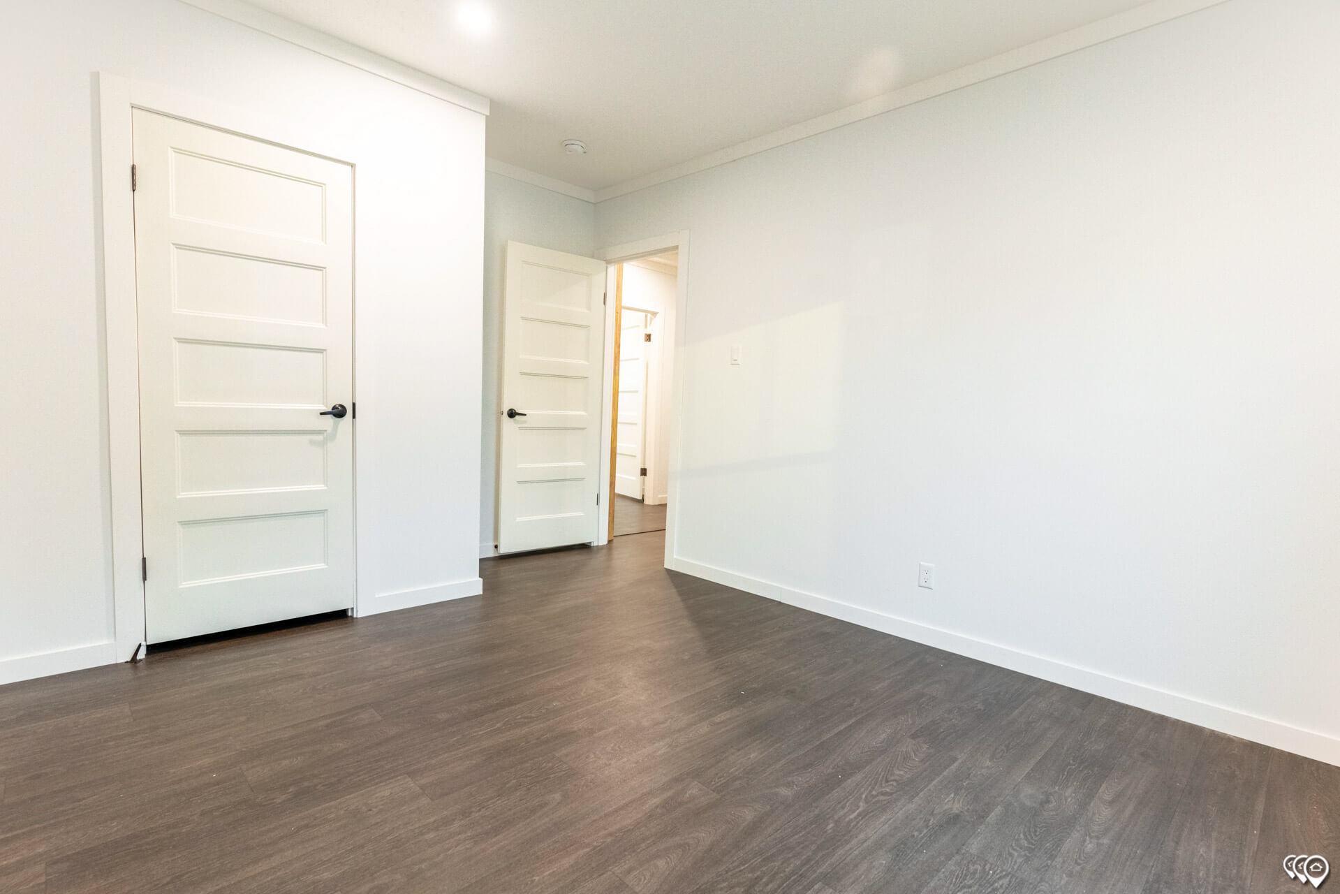 Empty room with light gray walls and dark wood laminate flooring. Two white paneled doors, one closed and one ajar, reveal a brightly lit hallway.