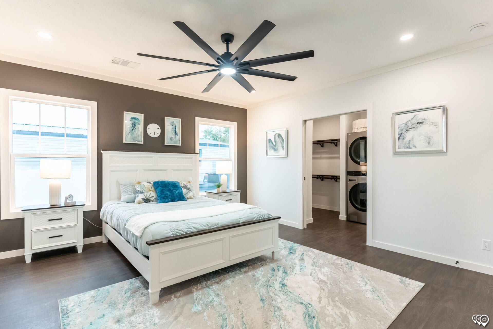 Modern bedroom with a white bed, teal accents, and a ceiling fan. There’s artwork on a gray accent wall, and a door opens to a laundry area. Cozy and serene ambiance.