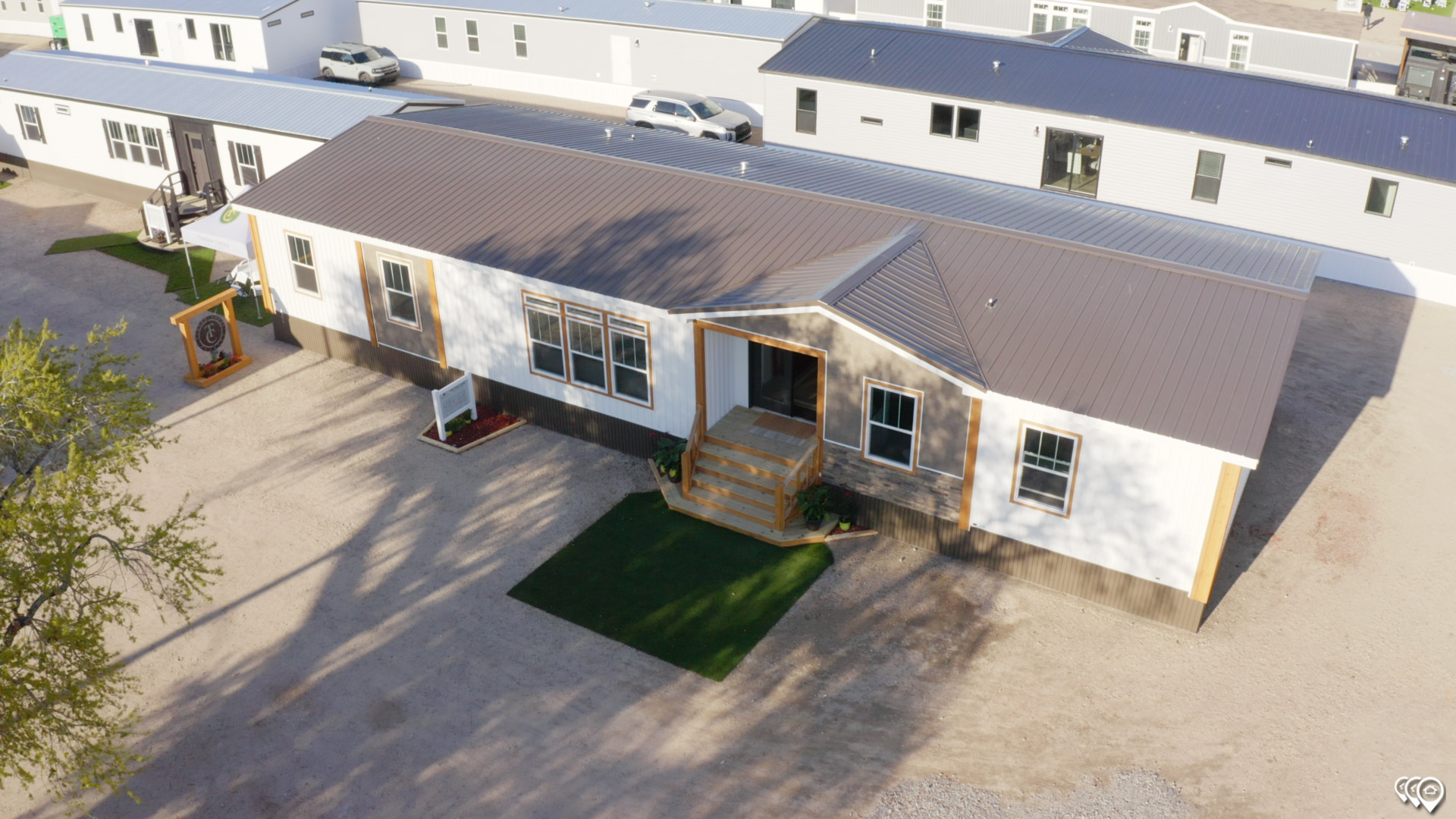 Aerial view of a modern, beige manufactured home with a sloped metal roof and white-trimmed windows. The home has a small, green lawn and steps leading to the entrance, surrounded by a gravel yard. Nearby, similar homes can be seen, with clear sunlight casting soft shadows, creating a calm suburban feel.
